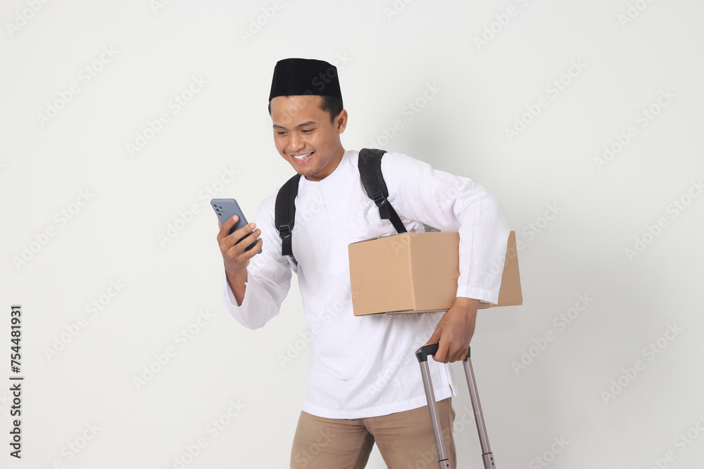 Portrait of happy Asian muslim man carrying cardboard box and suitcase while holding mobile phone. Going home for Eid Mubarak. Isolated image on white background Portrait of happy Asian muslim man carrying cardboard box and suitcase while holding mobile phone. Going home for Eid Mubarak. Isolated image on white background
