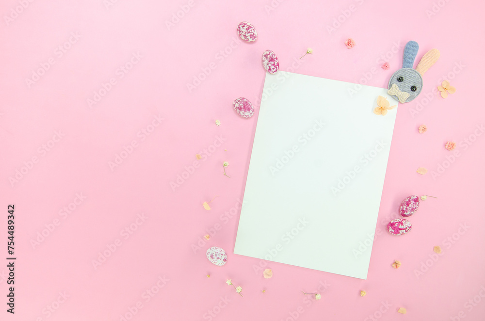 Easter party concept. Flat lay, top view of blank sheet of paper, easter bunny, multicolored candy eggs, small flowers on isolated pastel pink background