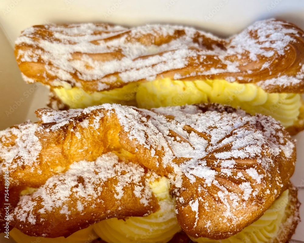 close in a cardboard box lie two eclairs with custard sprinkled with powdered sugar. side view, sweets close in a cardboard box lie two eclairs with custard sprinkled with powdered sugar. side view, sweets