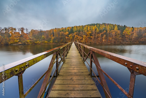 Beautiful view to long steel and wood bridge over big and silent lake with autumn golden trees and bushes around at cloudy and rainy morning