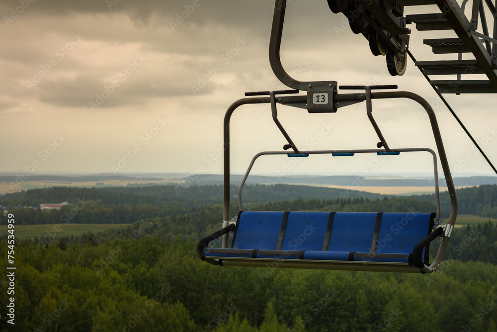 empty four-seater open cabin of cable car chairlift with blue seats moves over hilly forest against cloudy sky empty four-seater open cabin of cable car chairlift with blue seats moves over hilly forest against cloudy sky