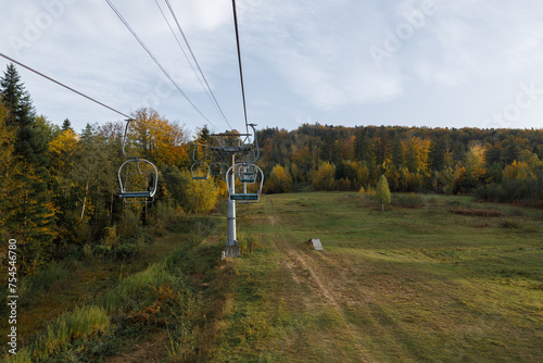 A ski lift ascends a forested hill under cloudy skies in outumn