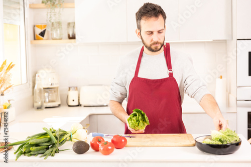 A man is preparing a salad in a kitchen. He is wearing a red apron and holding a head of lettuce. The kitchen is well-equipped with a toaster  a blender  and a microwave