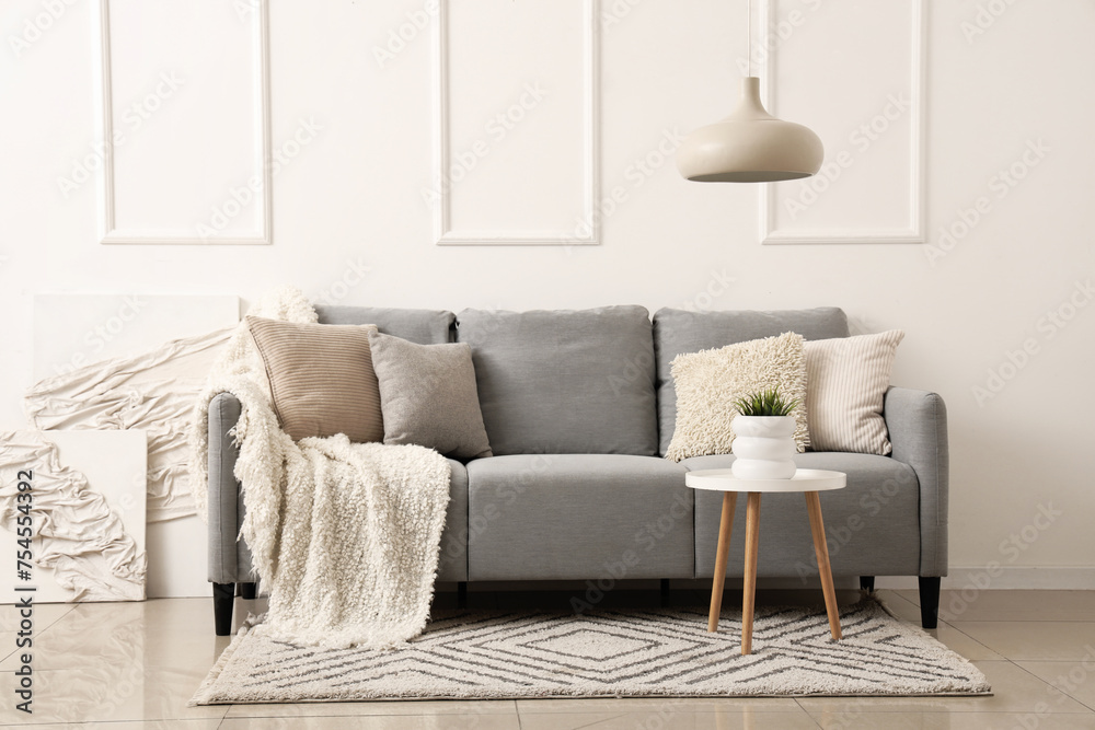 Stylish grey sofa with pillows, blanket and paintings near white wall - obrazy, fototapety, plakaty 