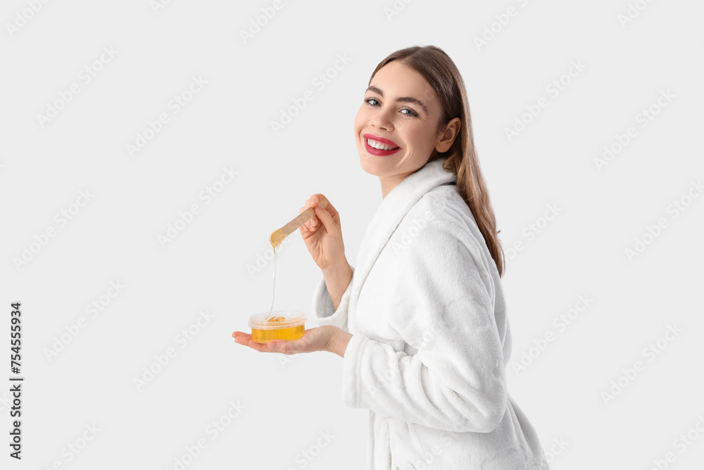 Beautiful young happy woman in bathrobe holding container with sugaring paste and spatula on grey background Beautiful young happy woman in bathrobe holding container with sugaring paste and spatula on grey background