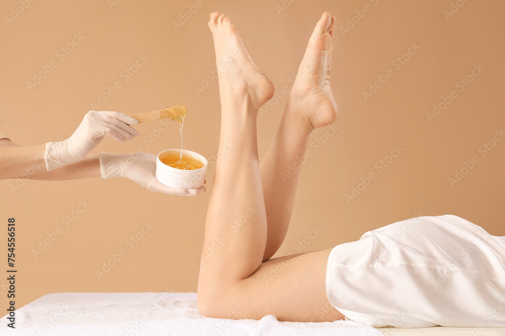 Naklejka premium Beautician epilating legs of beautiful young woman with sugaring paste on brown background