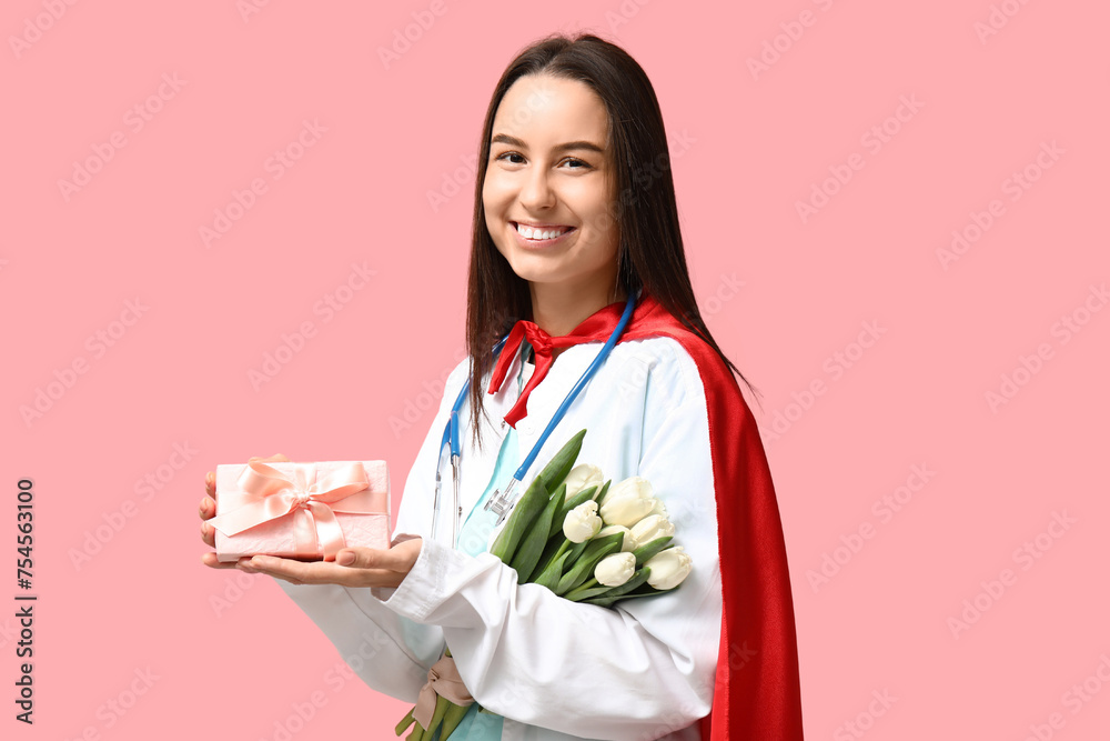 Female doctor with superhero cloak holding white tulips and gift box on pink background. Women's Day celebration Female doctor with superhero cloak holding white tulips and gift box on pink background. Women's Day celebration