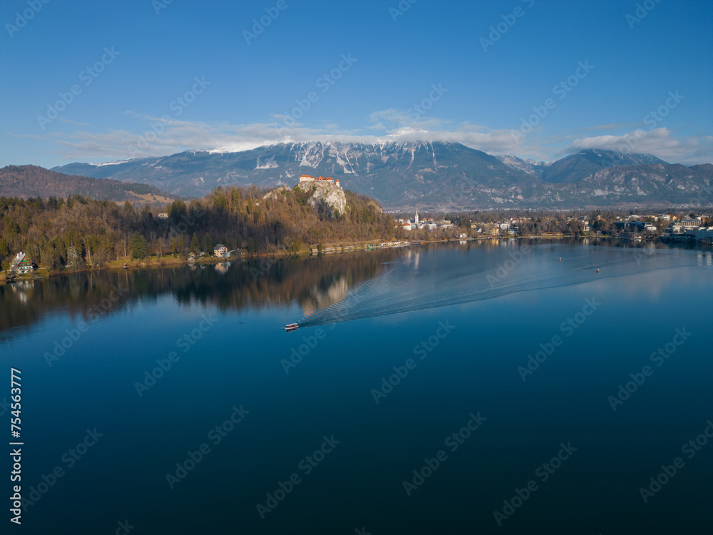 Aerial view of Lake Bled with the castle. Tourist boat sailing on a calm deep blue water surface with reflection. Panoramic view on a mountainous landscape in Slovenia. Snowless winter.