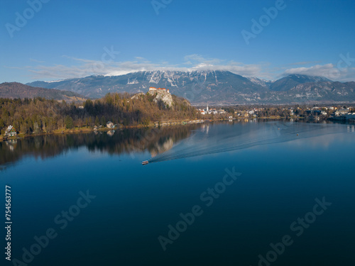 Aerial view of Lake Bled with the castle. Tourist boat sailing on a calm deep blue water surface with reflection. Panoramic view on a mountainous landscape in Slovenia. Snowless winter.
