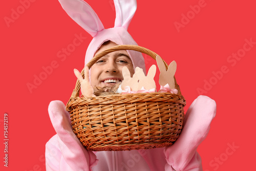 Happy young man in Easter bunny costume holding basket with gifts on red background