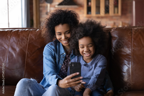 Happy African 6s girl and young mother sit on sofa with smartphone, enjoy video conference talk event with family, smiling watching funny online content, amusing videos. Modern technology, fun concept photo