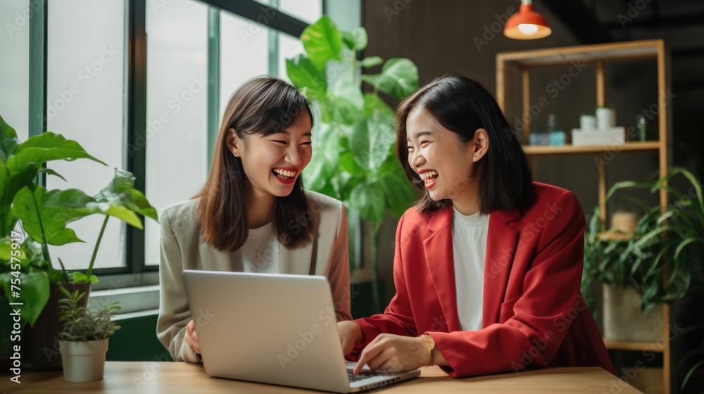 Two Asian women sitting in front of a laptop against office background with green plants.