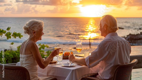 A senior couple sits at a table enjoying a meal and wine, with a beautiful ocean sunset in the background