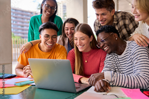 A large group of young multiracial students gathered in classroom smiling and having fun watching something funny on laptop. Generation z friends using and looking screen computer inside