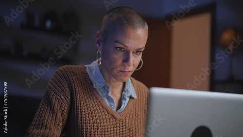 Shaved independent adult woman concentrating using laptop in living room at night. Serious Caucasian female looking for information on computer at home. Domestic life, electronic devices and leisure 