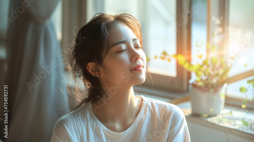 A young woman in a casual white shirt closes her eyes, relishing the warm sunlight by a window with blooming flowers.