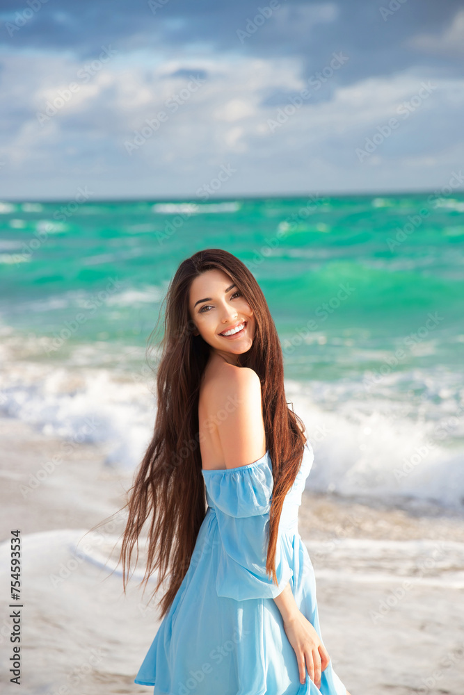 A beautiful brunette with long hair in a blue dress poses against the background of the ocean. Vacation A beautiful brunette with long hair in a blue dress poses against the background of the ocean. Vacation