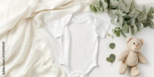 Cozy composition with a white baby bodysuit, teddy bear, and eucalyptus branch on a cream fabric background.