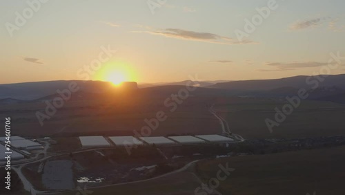 Aerial photography by drone over a large field with green and red grass. Mountains on the horizon during sunset. An empty asphalt road next to a vacant lot. One big cloud on a clear blue sky