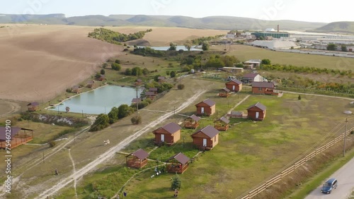 Old Believers village in a green field with a rectangular pond. Wooden houses on the shore of the lake. Aerial photography. High hills and mountains on the horizon. Asphalt road