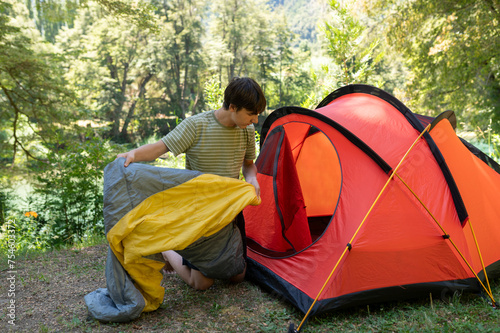 A young man arranges his camping tent and sleeping bag at the campsite during his summer walk through the mountains of southern Argentina. Surrounded by green, rivers and mountains.