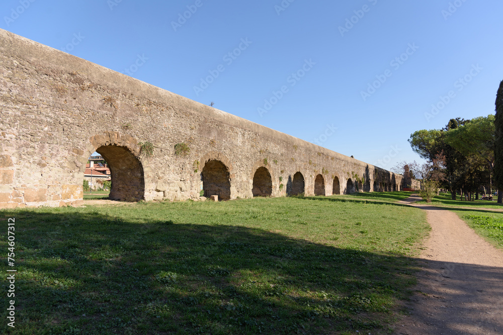 The long arched wall of an ancient Roman aqueduct along a walking path in a park, evening shadows from the setting sun shadows fall on the ground in a beautiful pattern