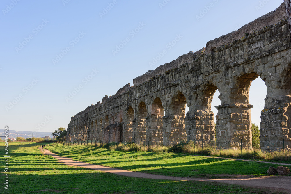 The long arched wall of an ancient Roman aqueduct along a walking path in a park, evening shadows from the setting sun shadows fall on the ground in a beautiful pattern