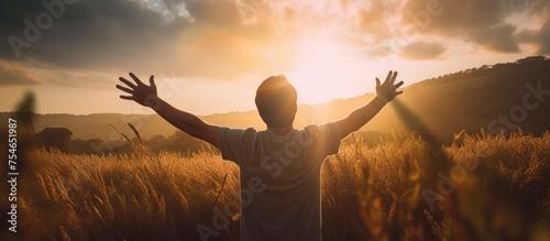 A young man is standing in a field during the evening, reaching his arms out towards the sunlight.