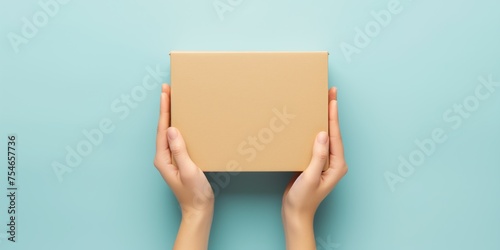 Top view of female hands holding a closed brown cardboard box on a vibrant teal background, representing delivery or packaging.