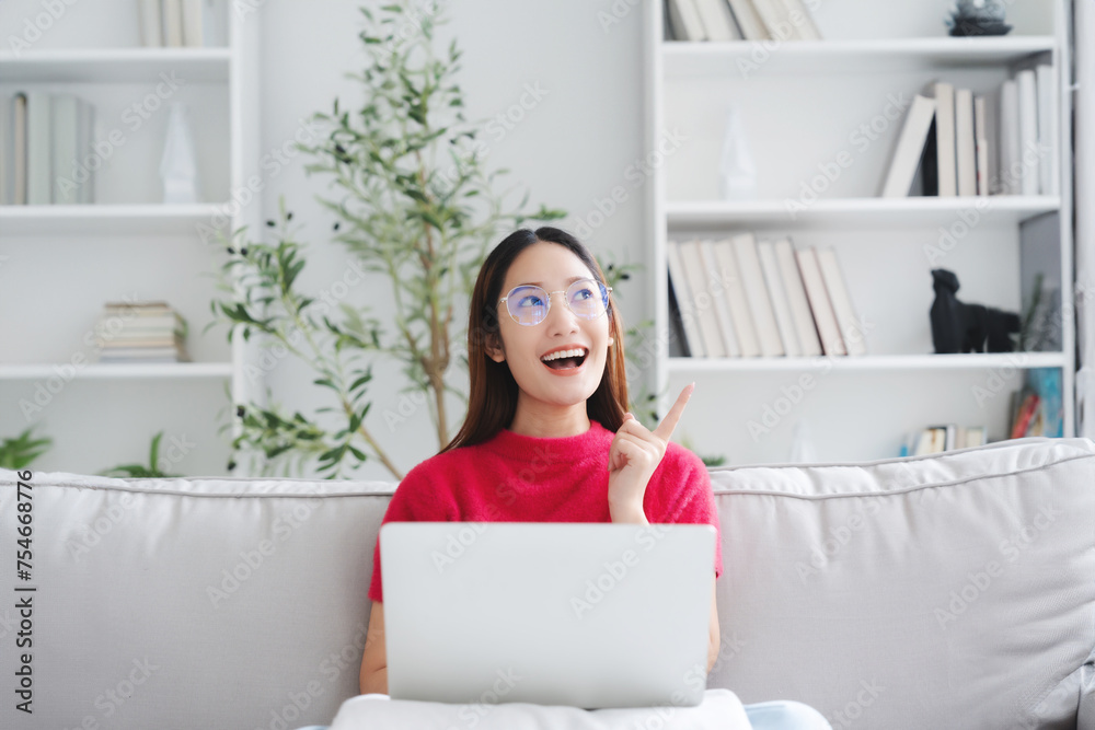 Happy young Asian woman wearing eyeglasses using laptop while seated on couch at home Happy young Asian woman wearing eyeglasses using laptop while seated on couch at home