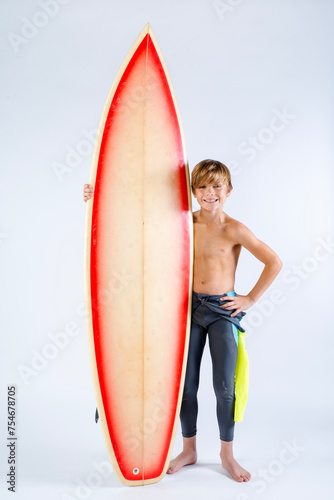 Smiling cute surfer boy with wet suit around his waist standing with his surfboard