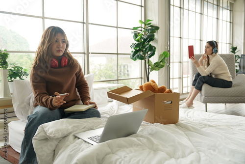 Foto Two Asian girls sitting on bed doing homework, working at home with laptops, just moved into a newly rented apartment, female rootmate sitting on sofa reading tablet