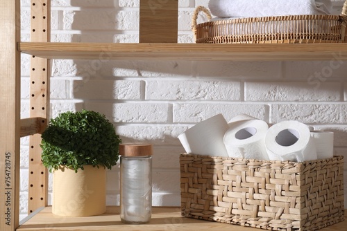 Toilet paper rolls in wicker basket, floral decor and cotton pads on wooden shelf against white brick wall