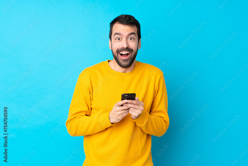 Young caucasian man over isolated blue background surprised and sending a message Young caucasian man over isolated blue background surprised and sending a message
