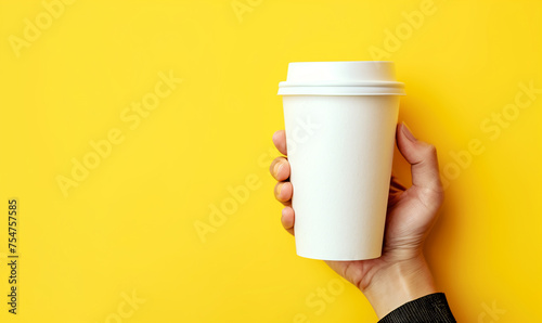 hand holding a take-away / to-go white cup on a yellow background with copy space, cup mockup 