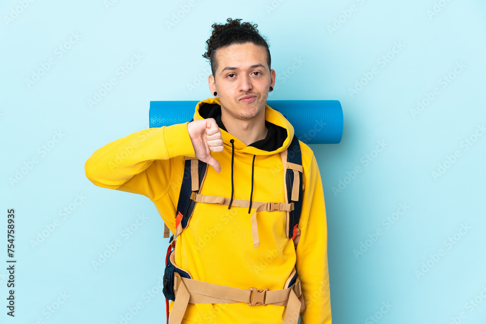 Young mountaineer man with a big backpack over isolated blue background showing thumb down sign