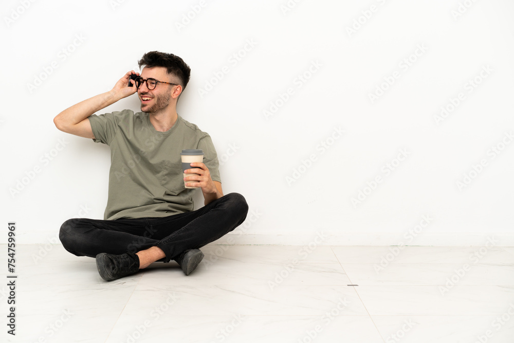 Young caucasian man sitting on the floor isolated on white background holding coffee to take away and a mobile Young caucasian man sitting on the floor isolated on white background holding coffee to take away and a mobile