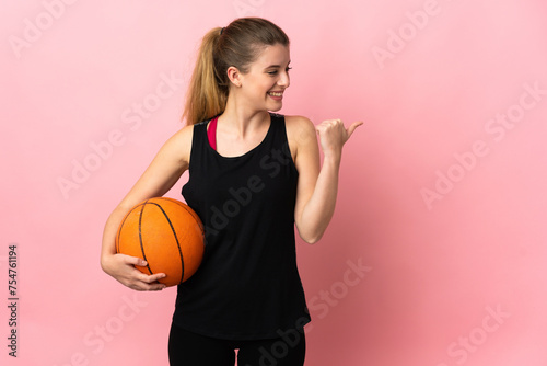 Young blonde woman playing basketball isolated on pink background pointing to the side to present a product