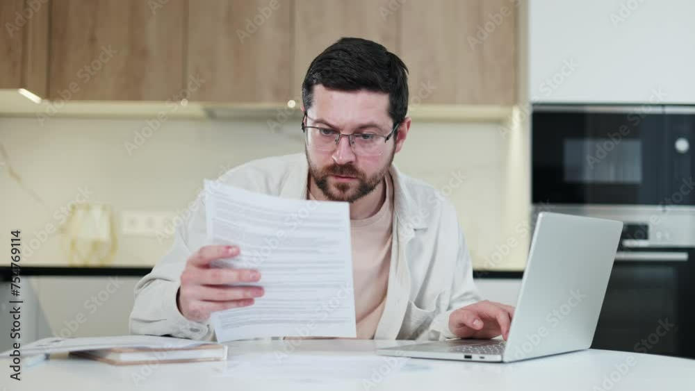 Overworked male reviewing important documents and looking at personal laptop during work process at home. Busy caucasian man engaging in distance work indoors. Concept of deadline and people.
