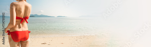 Panoramic banner image of sexy young woman or girl wearing a red bikini on a deserted tropical beach with a blue sky