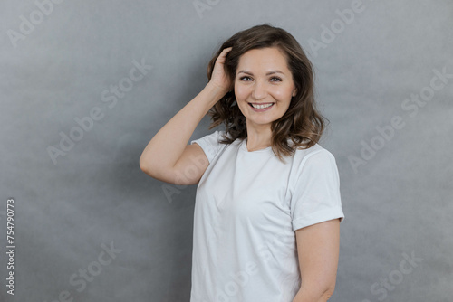 Portrait of cute young woman on grey background, holding hand near head, looking at camera and smiling, copy space.