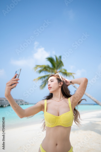 Woman in yellow bikini taking selfie near the coconut palm trees on tropical beach.