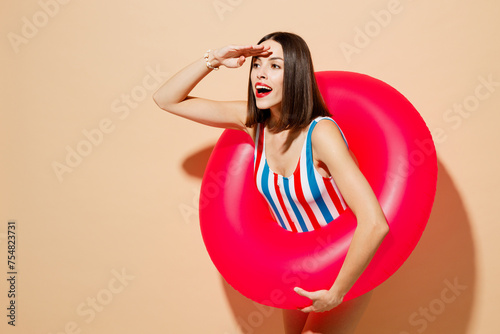 Side view young fun woman wear one-piece swimsuit near hotel pool hold pink inflatable rubber ring look far away distance isolated on plain beige background. Summer vacation sea rest sun tan concept.