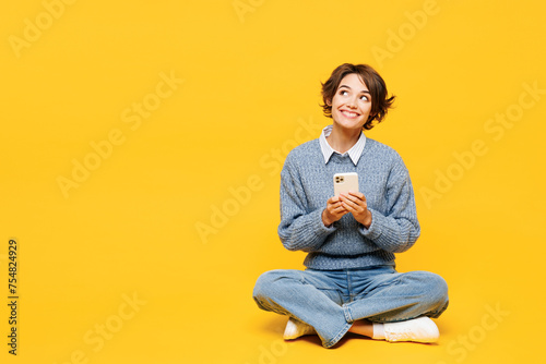Full body young woman wears grey knitted sweater shirt casual clothes sits hold in hand use mobile cell phone look aside on area isolated on plain yellow background studio portrait. Lifestyle concept.