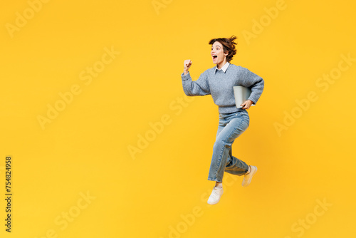 Full body side view young IT woman wearing grey knitted sweater shirt casual clothes jump high hold run with laptop pc computer isolated on plain yellow background studio portrait. Lifestyle concept.