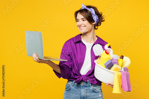 Young smart IT woman wear purple shirt hold basin with detergent bottles do housework tidy up use work on laptop pc computer surfing internet isolated on plain yellow background. Housekeeping concept.