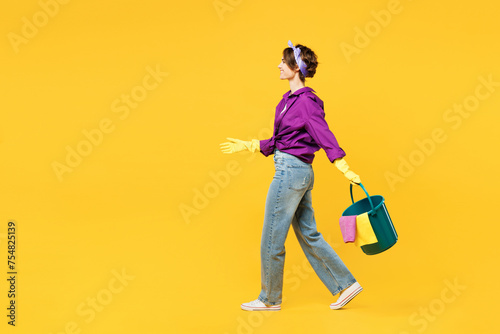 Full body side view young woman wear purple shirt casual clothes do housework tidy up walk go hold in hand bucket of water rag isolated on plain yellow background studio portrait Housekeeping concept