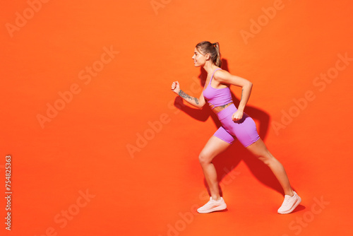 Full body sideways young fitness trainer instructor woman sportsman wears top shorts purple clothes train in home gym going to run isolated on plain orange background. Workout sport fit abs concept. © ViDi Studio