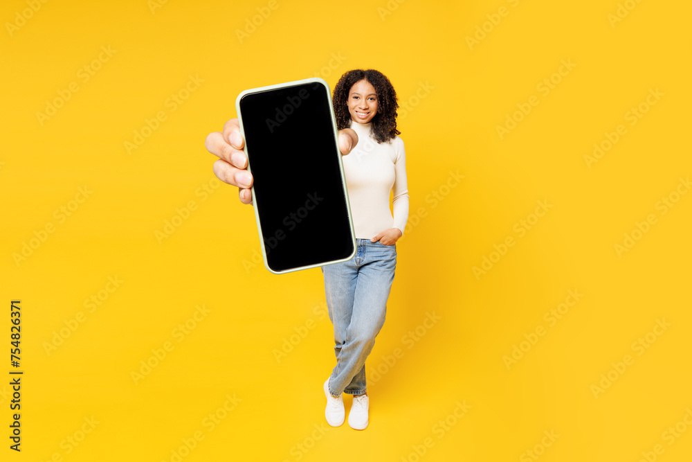 Full body little kid teen girl of African American ethnicity wear white casual clothes hold use close up blank screen mobile cell phone isolated on plain yellow background Childhood lifestyle concept Full body little kid teen girl of African American ethnicity wear white casual clothes hold use close up blank screen mobile cell phone isolated on plain yellow background Childhood lifestyle concept