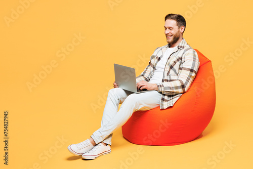 Full body young IT man wear brown shirt casual clothes sit in bag chair hold use work on laptop pc computer chat online isolated on plain yellow orange background studio portrait. Lifestyle concept photo